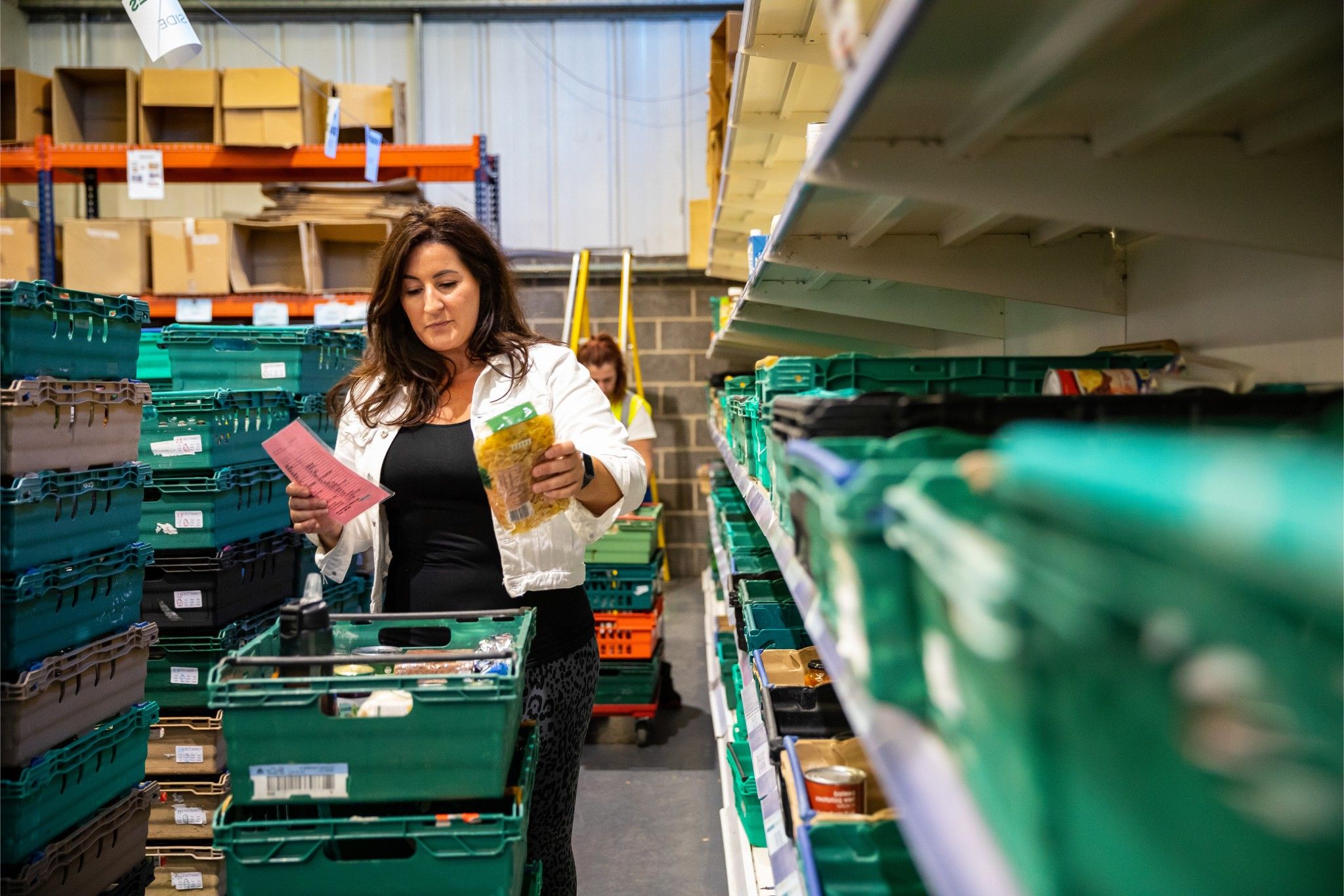 Lady in a white jacket is taking food items from the shelves and putting them in green crates in a warehouse.